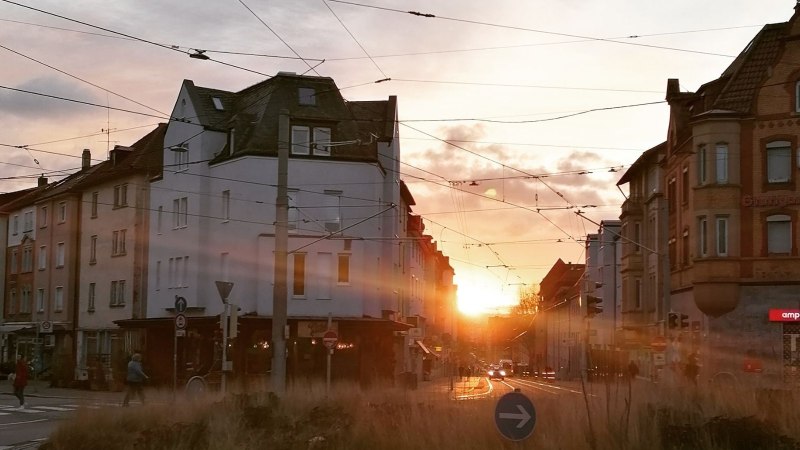 Sonnenuntergang in einer Stadtstraße mit historischen Gebäuden und Straßenbahnkabeln. Menschen überqueren die Straße, während die Sonne zwischen den Häusern untergeht., © SMG Sonnenuntergang in einer Stadtstraße mit historischen Gebäuden und Straßenbahnkabeln. Menschen überqueren die Straße, während die Sonne zwischen den Häusern untergeht., © SMG