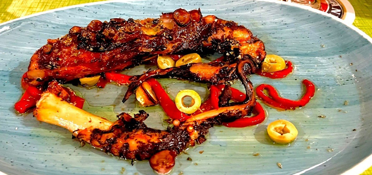 Gegrillter Oktopus mit roten Paprikastreifen und grünen Oliven auf einem blauen Teller angerichtet., © Restaurant Neckarau, P. Kotaidis