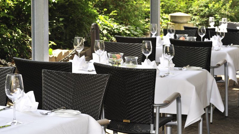 Elegante Restaurantterrasse mit gedeckten Tischen, wei&szlig;en Tischdecken und Weingl&auml;sern, umgeben von gr&uuml;ner Vegetation., &copy; Schupfnudl