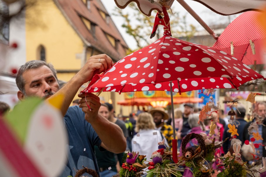 Ein Mann dekoriert einen roten Regenschirm mit weißen Punkten auf einem Kunsthandwerkermarkt. Im Hintergrund sind Besucher und bunte Stände zu sehen., © Stadt Nürtingen Ein Mann dekoriert einen roten Regenschirm mit weißen Punkten auf einem Kunsthandwerkermarkt. Im Hintergrund sind Besucher und bunte Stände zu sehen., © Stadt Nürtingen