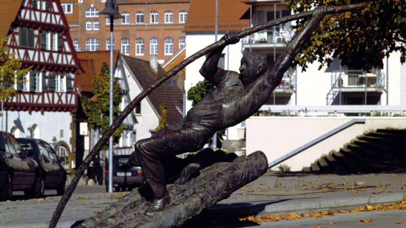 Bronzestatue eines Flößers, der mit einem langen Stab auf einem Baumstamm balanciert. Im Hintergrund Fachwerkhäuser und Herbstlaub auf dem Boden., © Stuttgart-Marketing GmbH