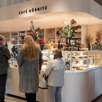 Theke mit Getr&auml;nken und Kuchenvitrine, dekoriert mit Pflanzen. Drei Personen stehen an., &copy; SMG, Sarah Schmid