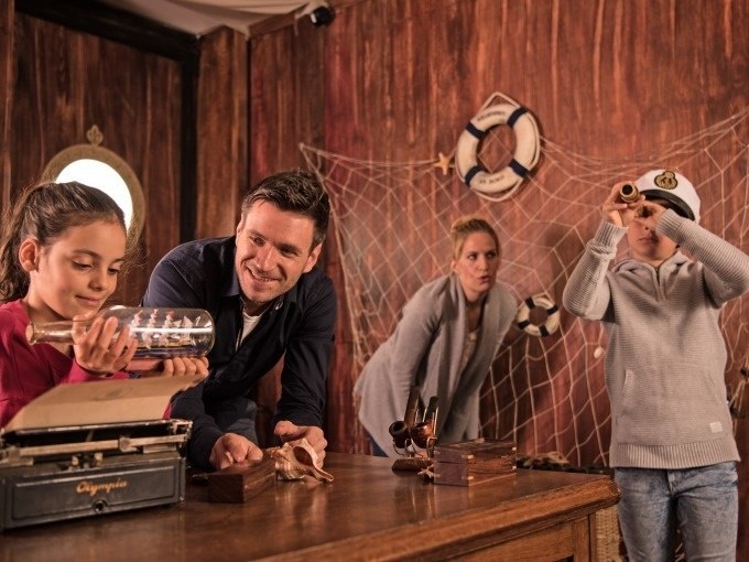 Familie in einem Escape Room mit Piratenthema. Ein Mädchen hält eine Flaschenpost, während ein Junge durch ein Fernrohr schaut. Im Hintergrund ein Fischernetz., © ExitGames Stuttgart Familie in einem Escape Room mit Piratenthema. Ein Mädchen hält eine Flaschenpost, während ein Junge durch ein Fernrohr schaut. Im Hintergrund ein Fischernetz., © ExitGames Stuttgart