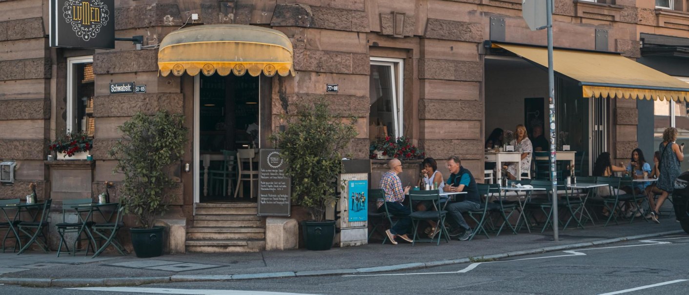 Café mit gelbem Vordach an einer Straßenecke. Gäste sitzen draußen an Tischen. Schild mit 'Lumen' und Straßenschild 'Schwabstr.' sichtbar., © SMG, Sarah Schmid Café mit gelbem Vordach an einer Straßenecke. Gäste sitzen draußen an Tischen. Schild mit 'Lumen' und Straßenschild 'Schwabstr.' sichtbar., © SMG, Sarah Schmid