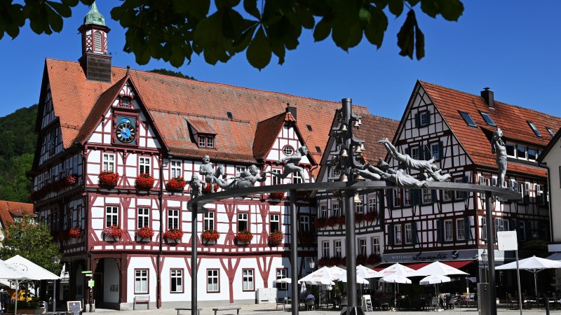 Fachwerkh&auml;user und eine Skulpturengruppe zieren den Marktplatz von Bad Urach. Die Geb&auml;ude sind mit roten Blumen geschm&uuml;ckt, der Himmel ist klar und blau., &copy; Bad Urach Tourismus