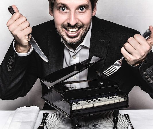 Ein Mann in Anzug h&auml;lt Messer und Gabel &uuml;ber einem kleinen schwarzen Klavier auf einem Teller. Er l&auml;chelt breit., &copy; Kulturhaus Schwanen