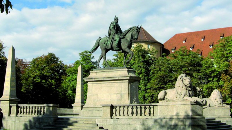 Reiterstatue auf dem Karlsplatz in Stuttgart, flankiert von Löwenstatuen und Obelisken, umgeben von Bäumen und einem historischen Gebäude im Hintergrund., © Stuttgart-Marketing GmbH