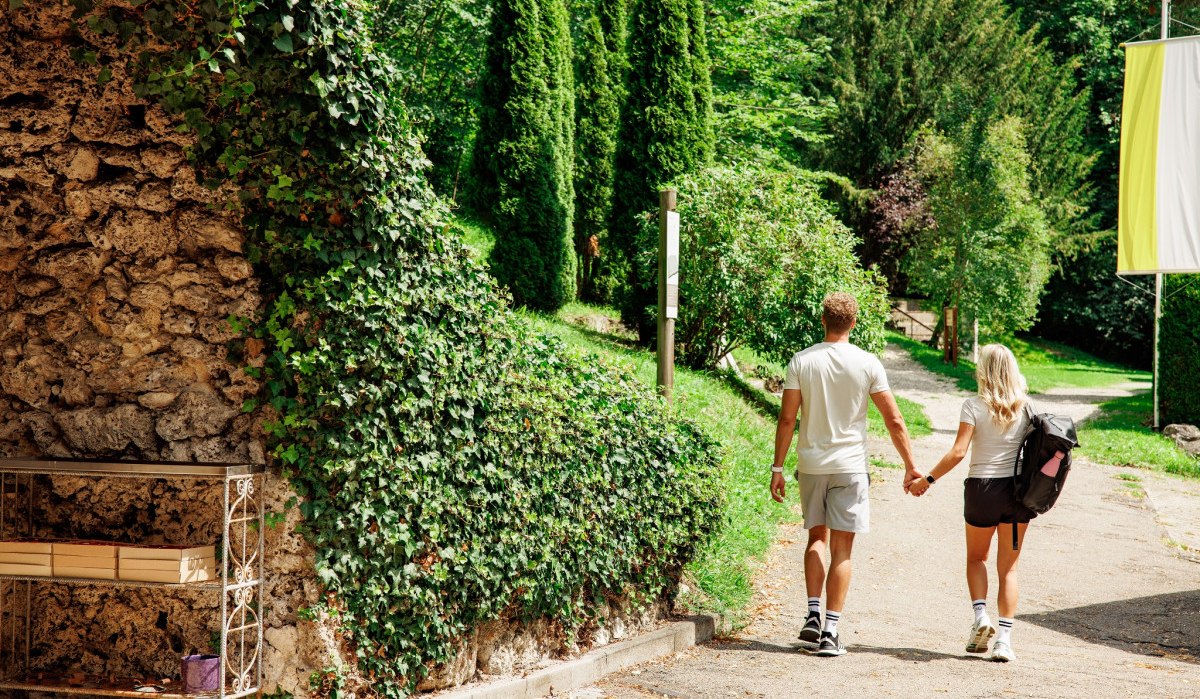 Ein Paar spaziert Hand in Hand auf einem Weg durch einen grünen, bewaldeten Park. Links ist eine mit Efeu bewachsene Steinwand., © Landkreis Göppingen
