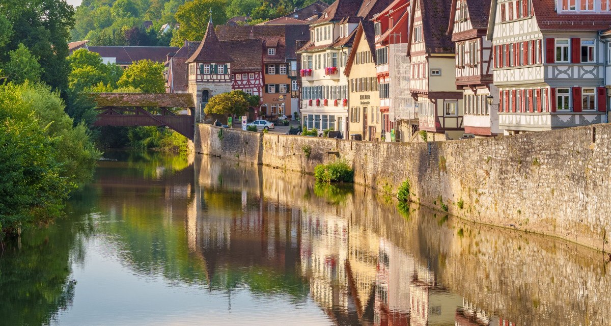 Fachwerkhäuser in Schwäbisch Hall spiegeln sich im Fluss. Eine Brücke und grüne Hügel im Hintergrund vervollständigen die malerische Szene., © Michael Kühneisen Fachwerkhäuser in Schwäbisch Hall spiegeln sich im Fluss. Eine Brücke und grüne Hügel im Hintergrund vervollständigen die malerische Szene., © Michael Kühneisen