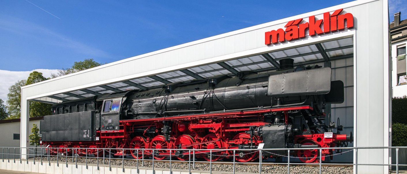 Eine schwarze Dampflokomotive mit roten Rädern steht vor dem Märklineum in Göppingen unter einem modernen Glasdach., © SMG, Sarah Schmid Eine schwarze Dampflokomotive mit roten Rädern steht vor dem Märklineum in Göppingen unter einem modernen Glasdach., © SMG, Sarah Schmid