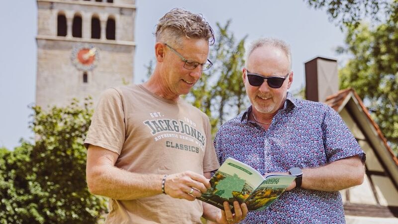 Zwei Männer stehen im Freien und schauen in ein Buch. Im Hintergrund ist ein Kirchturm zu sehen. Die Szene wirkt entspannt und frühlingshaft., © Wirtschaftsförderung Sindelfingen GmbH Zwei Männer stehen im Freien und schauen in ein Buch. Im Hintergrund ist ein Kirchturm zu sehen. Die Szene wirkt entspannt und frühlingshaft., © Wirtschaftsförderung Sindelfingen GmbH