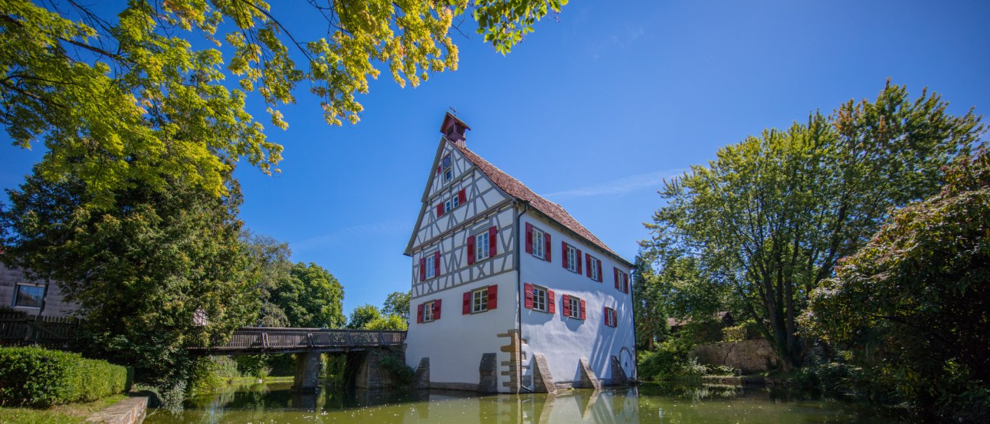 Fachwerkhaus mit roten Fensterläden an einem Teich, umgeben von Bäumen und einer Brücke im Hintergrund, unter blauem Himmel., © SMG, Achim Mende