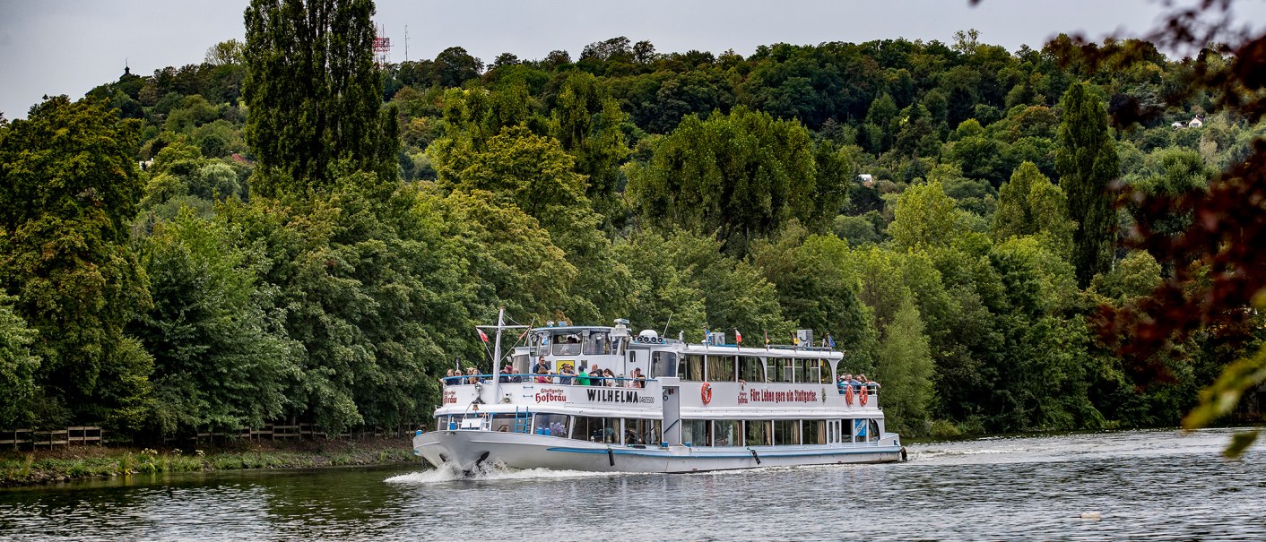 Das Ausflugsschiff Wilhelma fährt auf einem Fluss, umgeben von dichtem Wald und grünen Hügeln., © all copyrights are reserved by maks richter Das Ausflugsschiff Wilhelma fährt auf einem Fluss, umgeben von dichtem Wald und grünen Hügeln., © all copyrights are reserved by maks richter