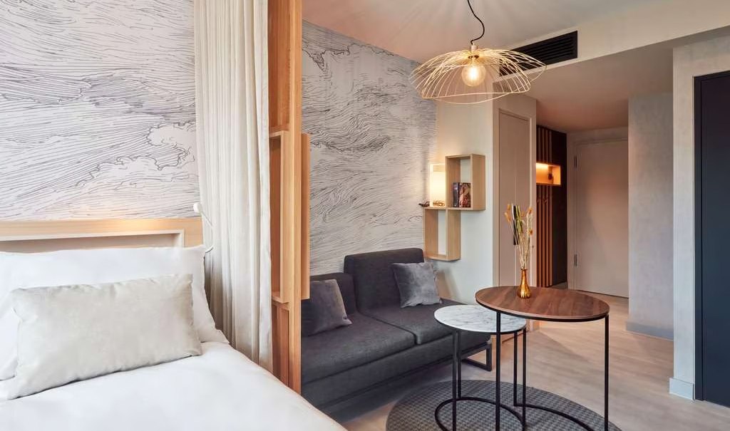 Modernes Studio mit Bett, Sofa, rundem Tisch und stilvoller Lampe. Wand mit abstraktem Muster, helle Farben und minimalistische Einrichtung., © Adagio Modernes Studio mit Bett, Sofa, rundem Tisch und stilvoller Lampe. Wand mit abstraktem Muster, helle Farben und minimalistische Einrichtung., © Adagio