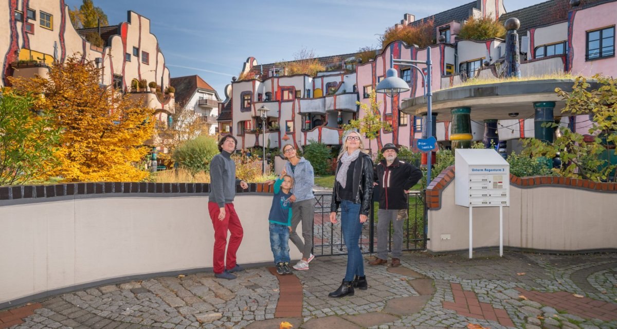 Menschen stehen vor einem bunten Hundertwasser-Gebäude mit unregelmäßigen Formen und begrünten Dächern., © Bildergalerie Attila Menschen stehen vor einem bunten Hundertwasser-Gebäude mit unregelmäßigen Formen und begrünten Dächern., © Bildergalerie Attila