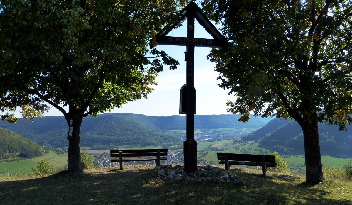 Holzkreuz zwischen zwei Bäumen, flankiert von Bänken, mit Blick auf ein grünes Tal und Hügel im Hintergrund., © ESA Holzkreuz zwischen zwei Bäumen, flankiert von Bänken, mit Blick auf ein grünes Tal und Hügel im Hintergrund., © ESA