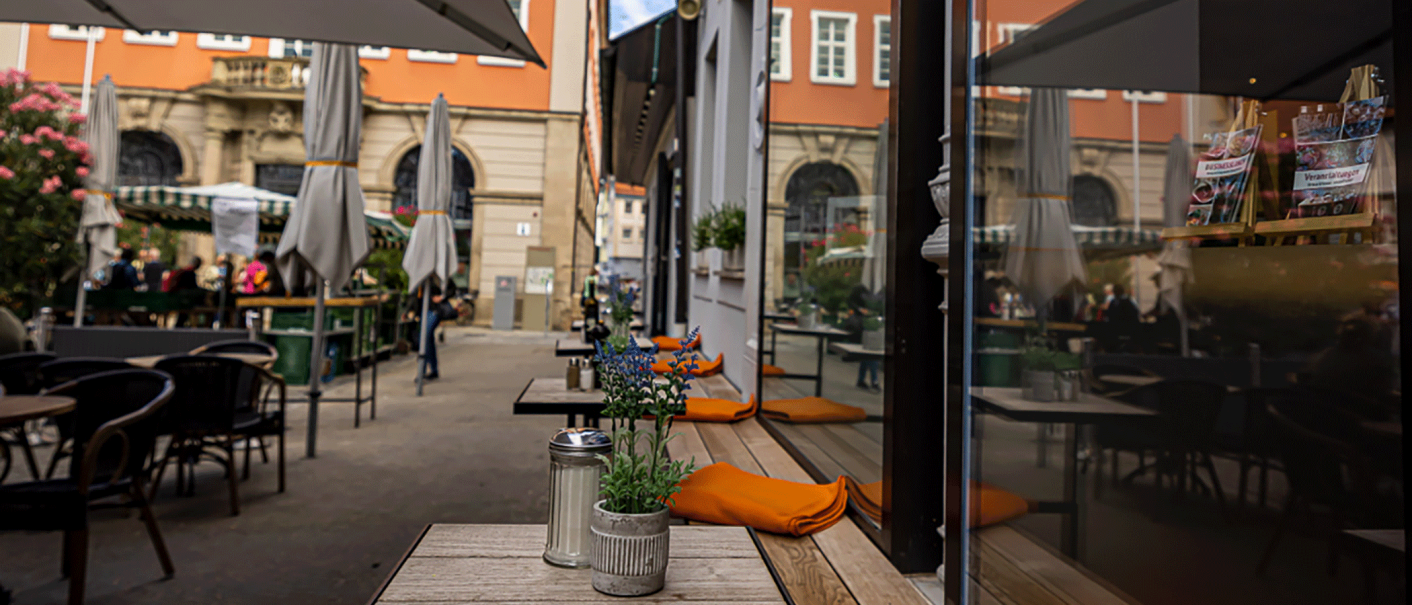 Ein gemütliches Straßencafé mit Tischen, Stühlen und Pflanzen. Im Hintergrund sind historische Gebäude und Menschen zu sehen., © Ama Deli Ein gemütliches Straßencafé mit Tischen, Stühlen und Pflanzen. Im Hintergrund sind historische Gebäude und Menschen zu sehen., © Ama Deli