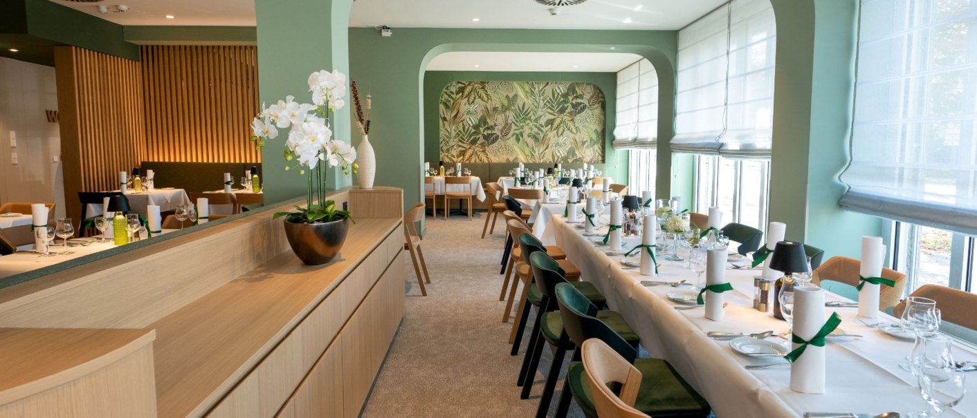 Eleganter Frühstücksraum mit grünen Wänden, gedeckten Tischen und Orchideen. Große Fenster lassen viel Licht herein., © Erikson Hotel Eleganter Frühstücksraum mit grünen Wänden, gedeckten Tischen und Orchideen. Große Fenster lassen viel Licht herein., © Erikson Hotel