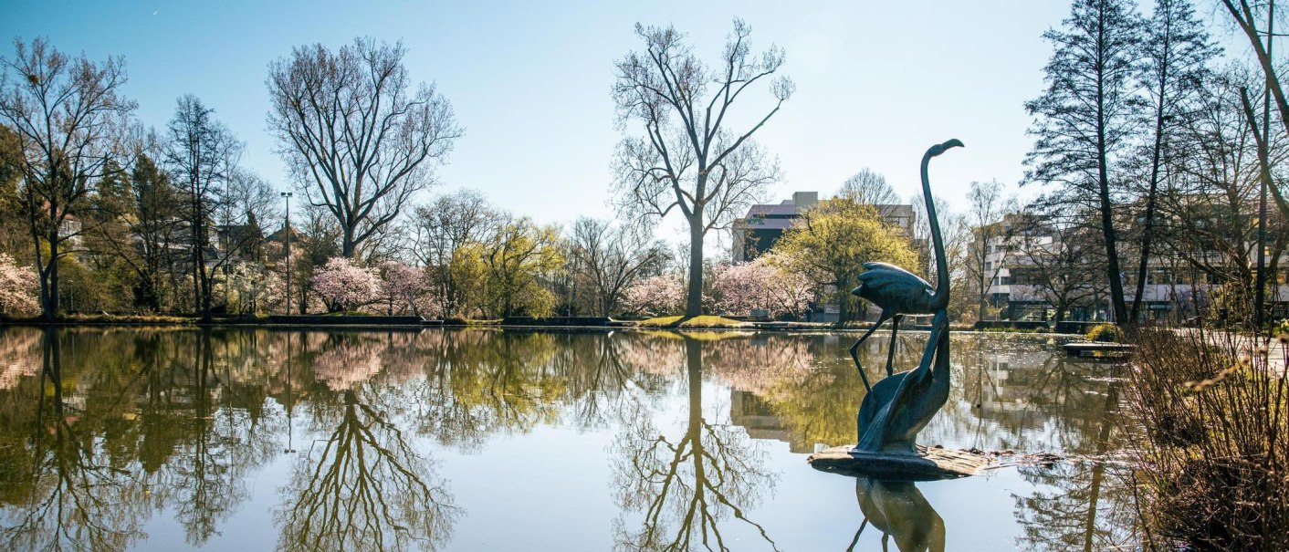 Ein See mit einer Vogelstatue im Vordergrund, umgeben von blühenden Bäumen und reflektierenden Wasserflächen unter klarem Himmel., © Stuttgart-Marketing GmbH, Sarah Schmid