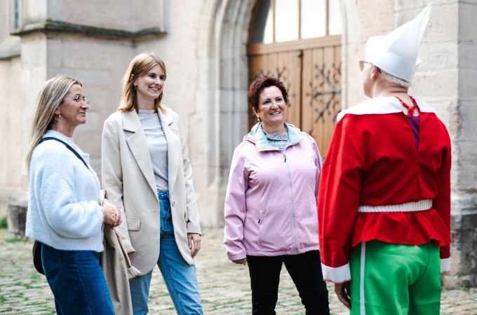 Drei Frauen unterhalten sich mit einer Person in einem bunten Kost&uuml;m vor einem historischen Geb&auml;ude. Die Szene wirkt fr&ouml;hlich und entspannt., &copy; Stadt N&uuml;rtingen