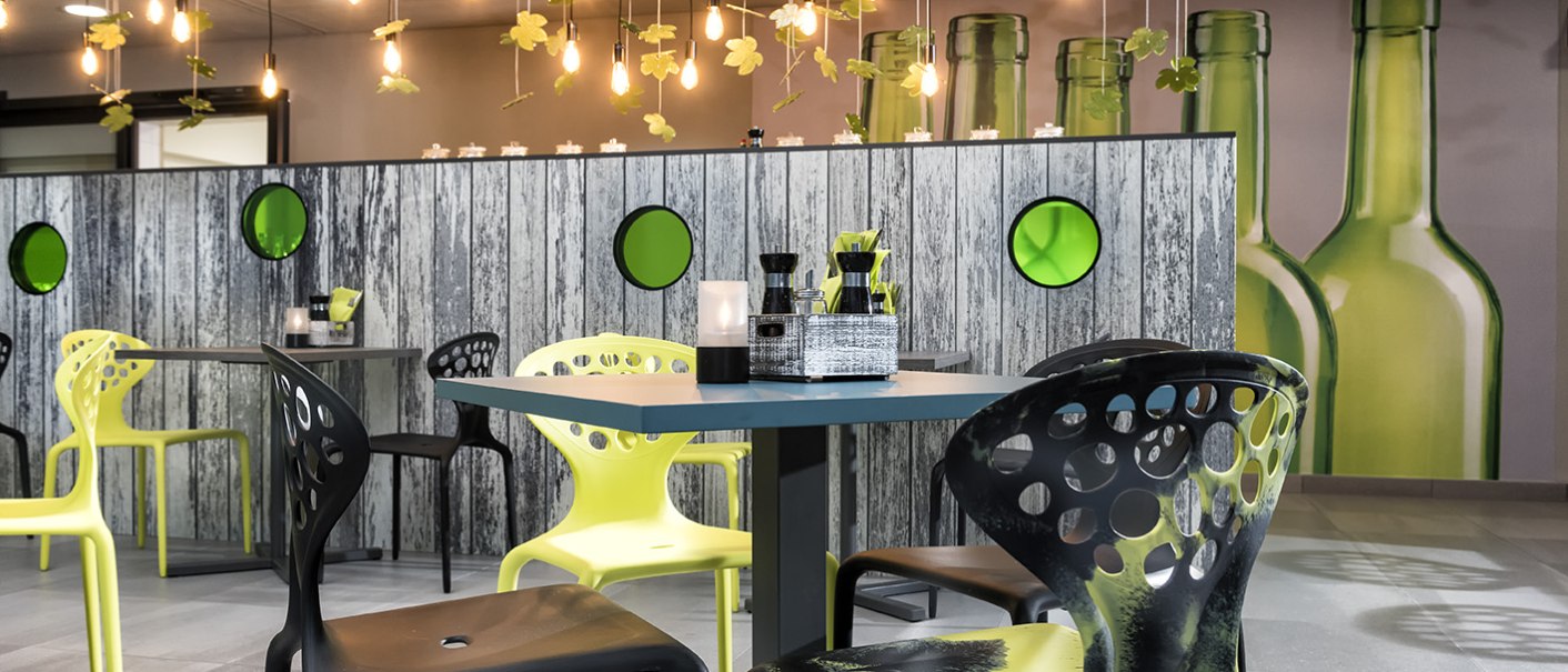 Modernes Restaurant mit gelben und schwarzen Stühlen, hängenden Flaschen und Blumen als Dekoration. Holzwand mit grünen Kreisen im Hintergrund., © ibis Styles Stuttgart Vaihingen