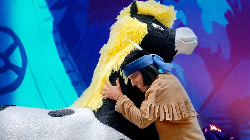 Ein Schauspieler in einem Yakari-Kost&uuml;m umarmt ein gro&szlig;es Pl&uuml;schpferd. Der Hintergrund ist bunt und zeigt eine animierte Szene., &copy; Whynot Events UG