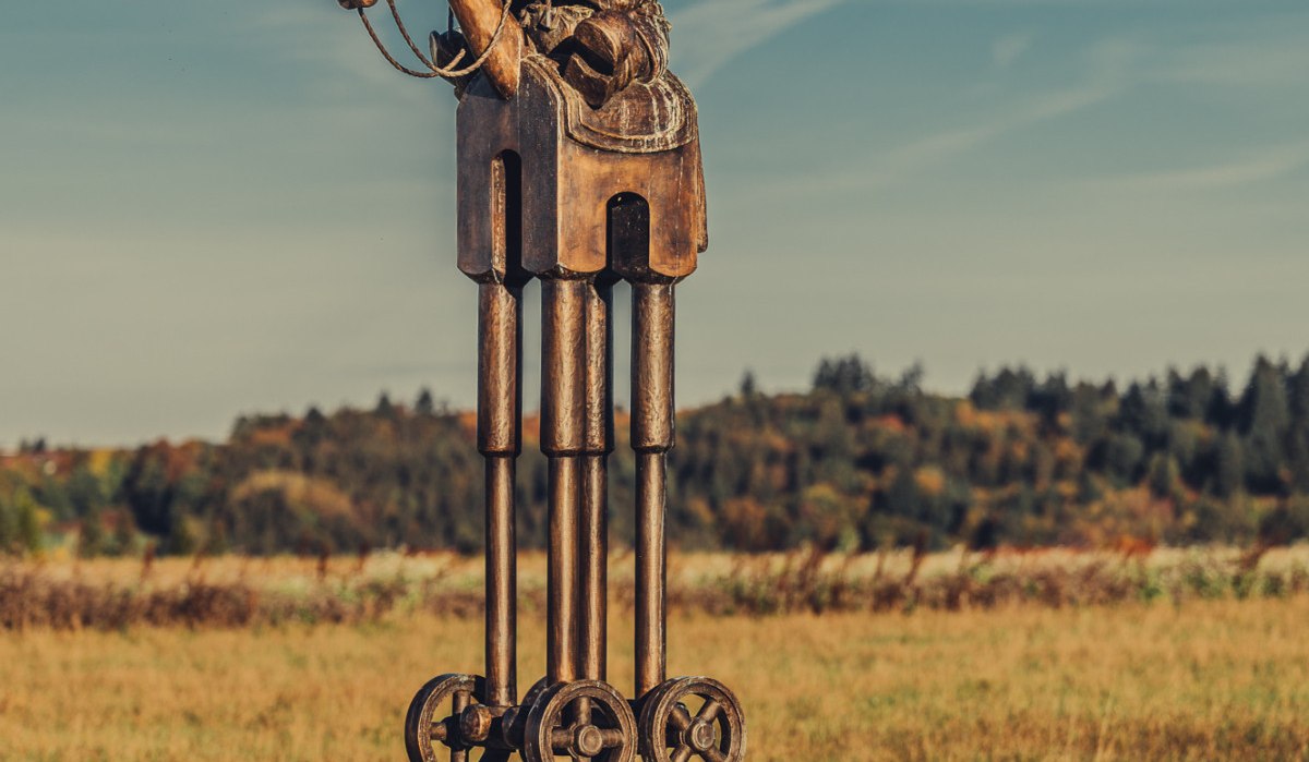 Metallskulptur eines Reiters auf einem Pferd mit Rädern, auf einem Sockel in einer Wiese unter blauem Himmel., © Landratsamt Ostalbkreis