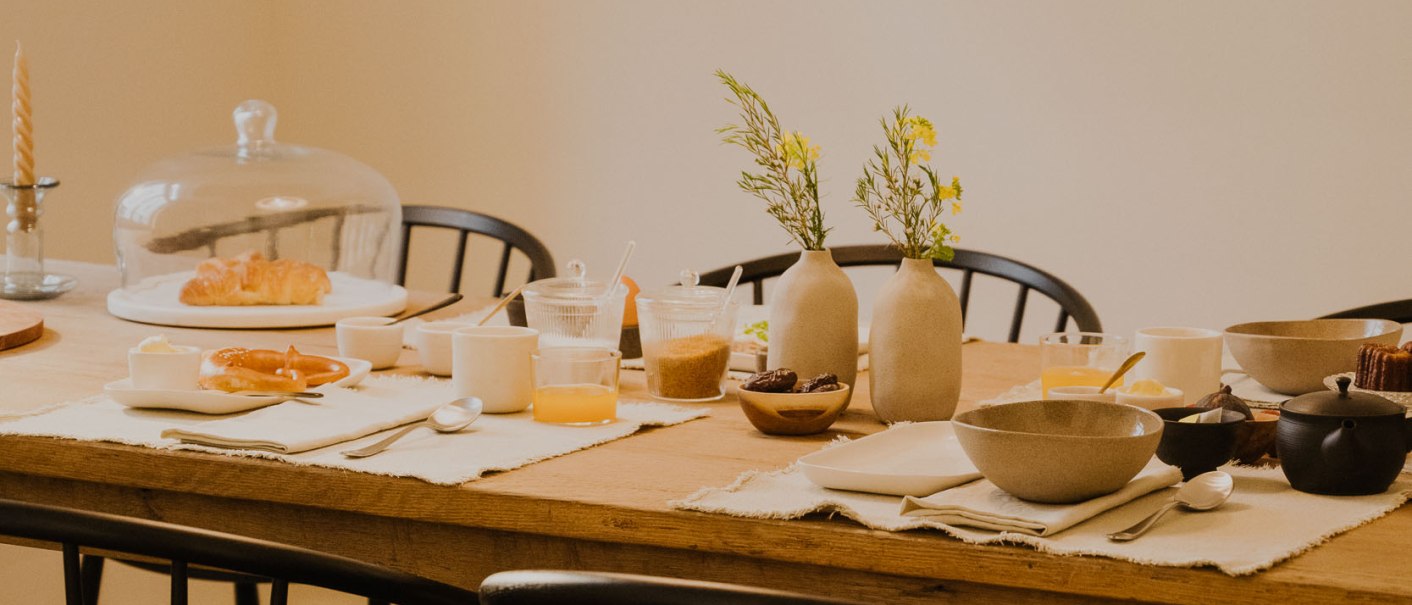 Ein Frühstückstisch mit Croissants, Saft, Geschirr und Blumenvasen. Eine Hängelampe beleuchtet den Tisch. Schwarze Stühle umgeben den Holztisch., © Peter Oliver Wolff / DAS EDITH