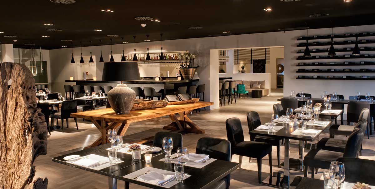 Modernes Restaurant mit eleganter Einrichtung, gedeckten Tischen und stilvoller Beleuchtung. Im Hintergrund eine Bar mit Flaschenregal., © Wyndham Stuttgart Airport Messe