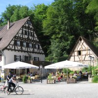 Ein Fachwerkhaus mit Biergarten, umgeben von Bäumen. Menschen sitzen an Tischen, während zwei Radfahrer im Vordergrund stehen., © Natur.Nah. Schönbuch & Heckengäu