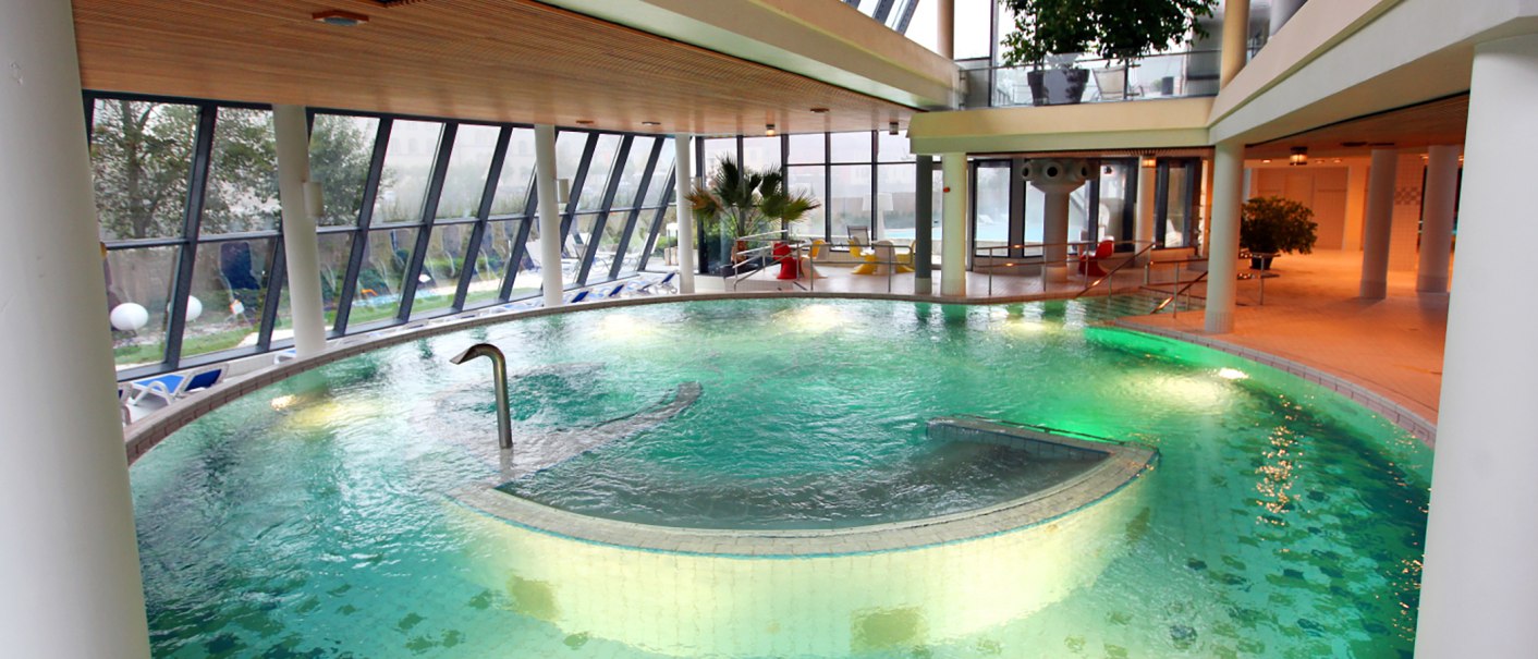 Modernes Schwimmbad mit großen Fenstern, grünem Wasser und Liegestühlen. Innenbereich mit Pflanzen und Sitzgelegenheiten., © Ufuk Arslan