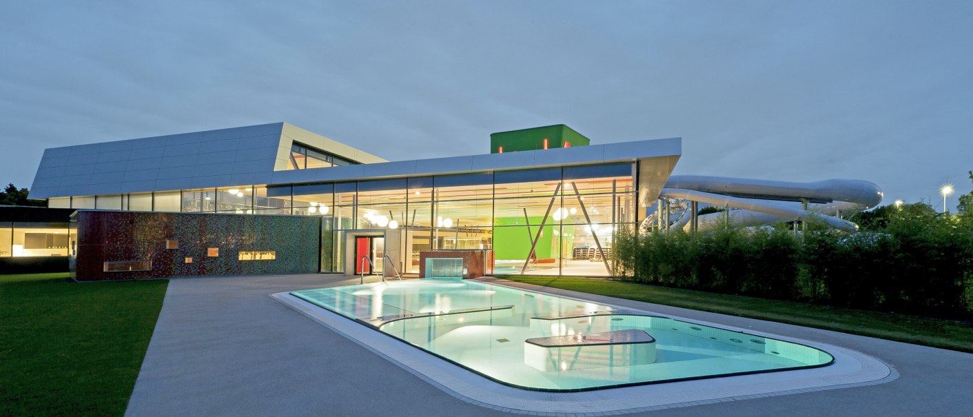 Modernes Schwimmbad mit beleuchtetem Außenpool und Glasfassade bei Dämmerung., © David Matthiessen Fotografie