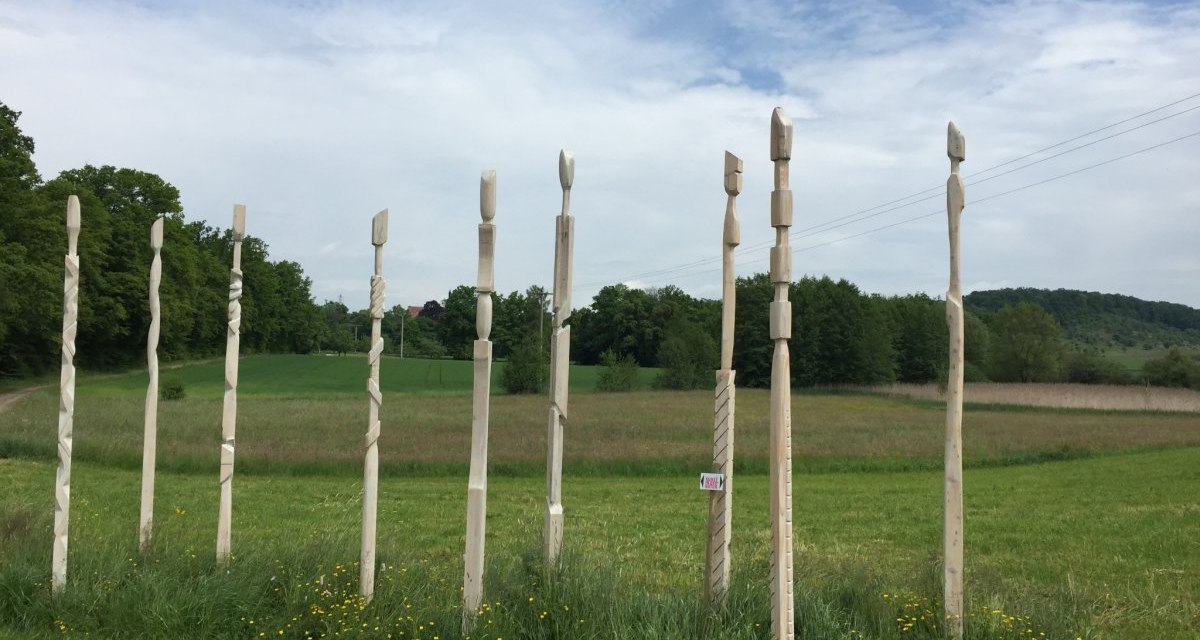 Holzskulpturen stehen in einer Reihe auf einer gr&uuml;nen Wiese. Im Hintergrund sind B&auml;ume und ein bewaldeter H&uuml;gel zu sehen., &copy; www.pro-cycl.de