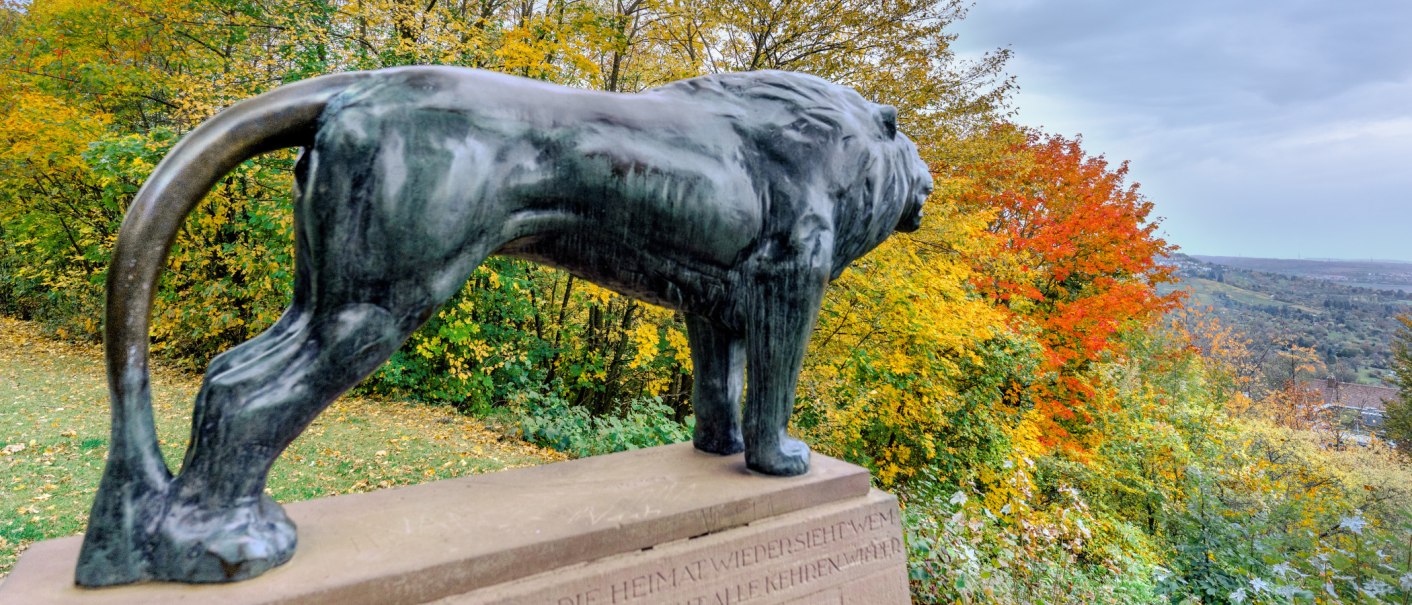 Bronzestatue eines Löwen auf einem Sockel, umgeben von buntem Herbstlaub und Blick auf eine weite Landschaft., © SMG, Martina Denker Bronzestatue eines Löwen auf einem Sockel, umgeben von buntem Herbstlaub und Blick auf eine weite Landschaft., © SMG, Martina Denker