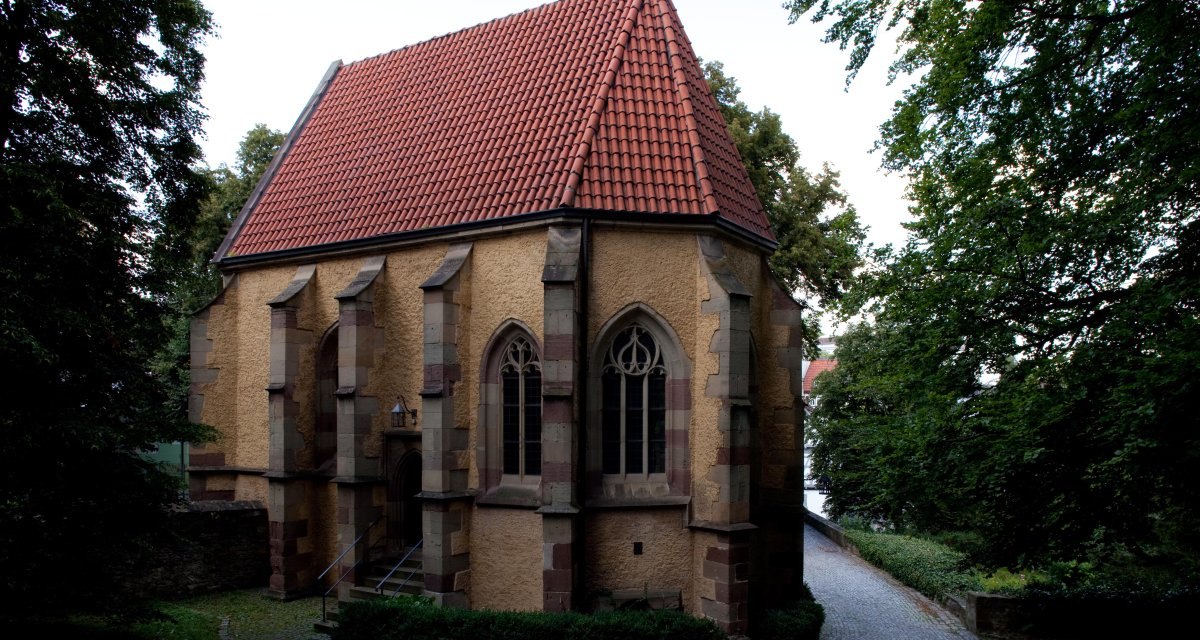 Kleine Kirche mit rotem Ziegeldach und gotischen Fenstern, umgeben von Bäumen und einem gepflasterten Weg., © Waiblingen - Stuttgart-Marketing GmbH Kleine Kirche mit rotem Ziegeldach und gotischen Fenstern, umgeben von Bäumen und einem gepflasterten Weg., © Waiblingen - Stuttgart-Marketing GmbH