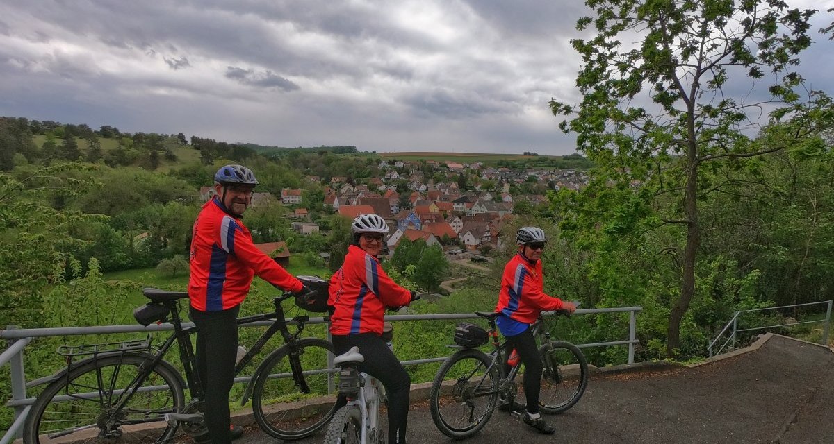 Drei Radfahrer in roten Jacken stehen auf einem Hügel und blicken auf ein Dorf im Tal. Der Himmel ist bewölkt, die Landschaft grün., © Land der 1000 Hügel - Kraichgau-Stromberg Drei Radfahrer in roten Jacken stehen auf einem Hügel und blicken auf ein Dorf im Tal. Der Himmel ist bewölkt, die Landschaft grün., © Land der 1000 Hügel - Kraichgau-Stromberg