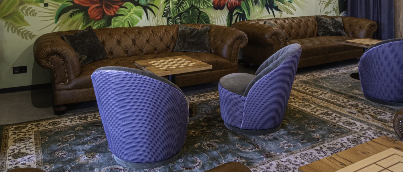 Gemütliche Sitzecke mit braunen Ledersofas, blauen Sesseln und einem Schachbrett-Tisch vor einer Wand mit tropischem Pflanzenmuster., © 13 6 Design GmbH (Judenhof 10, 89073 Ulm)