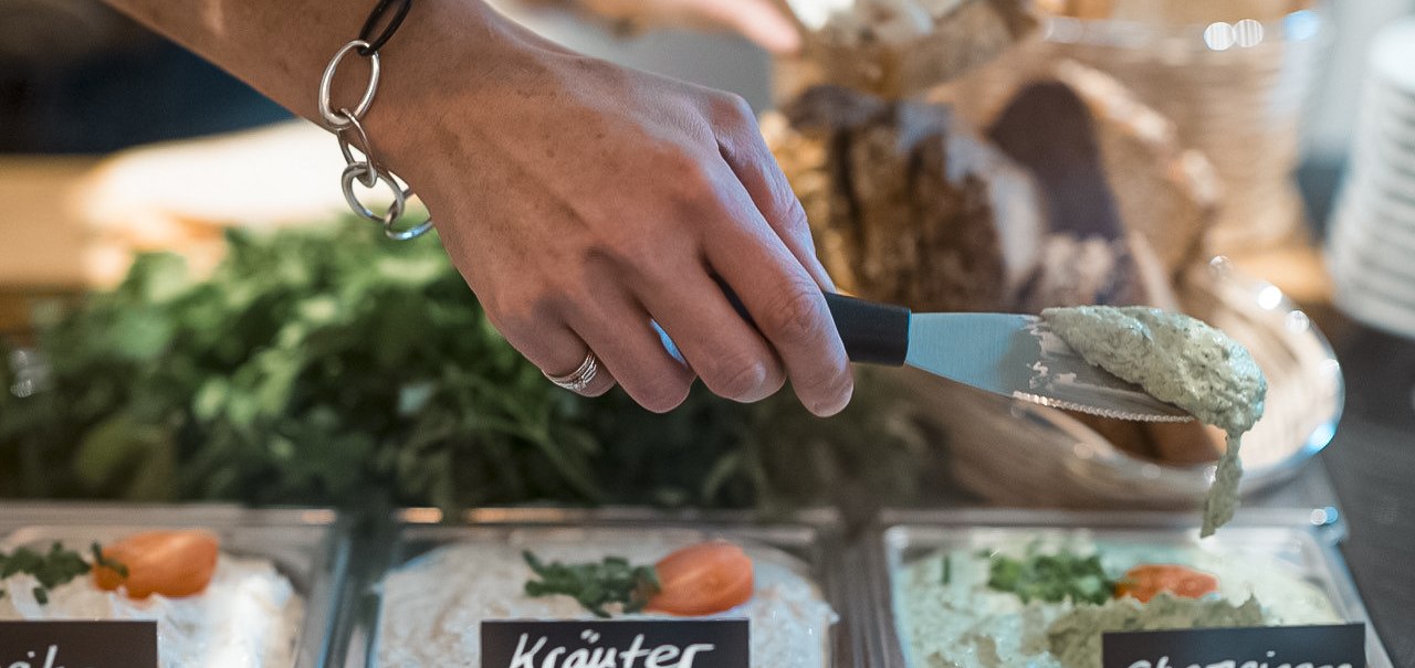 Eine Hand mit Armband nimmt Champignoncreme aus einer Schale. Daneben stehen Schalen mit Kräuterfrischkäse und Paprikacreme, im Hintergrund Brot., © Café Justus, Stuttgart