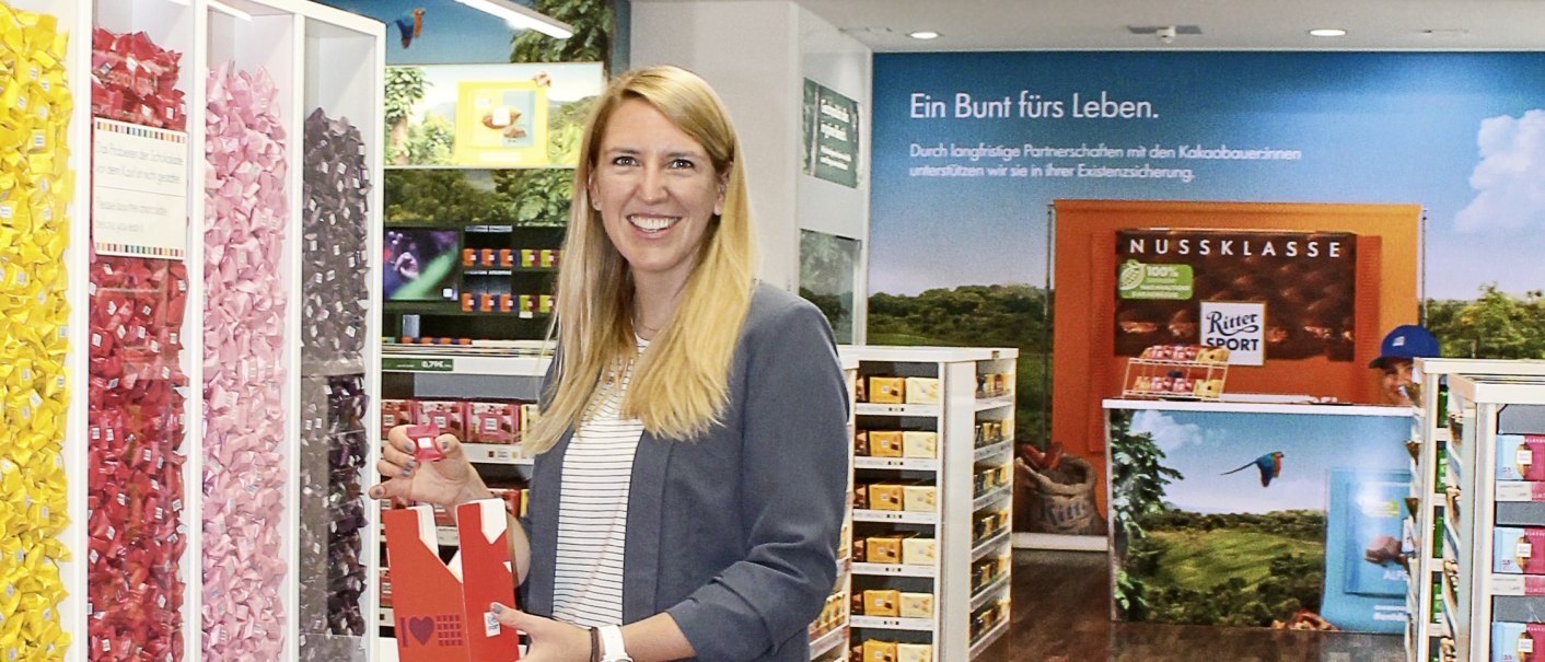 Eine Frau steht im Ritter Sport Schokoshop und hält eine Schachtel Schokolade. Im Hintergrund sind bunte Schokoladenregale zu sehen., © Alfred Ritter GmbH & Co. KG Eine Frau steht im Ritter Sport Schokoshop und hält eine Schachtel Schokolade. Im Hintergrund sind bunte Schokoladenregale zu sehen., © Alfred Ritter GmbH & Co. KG