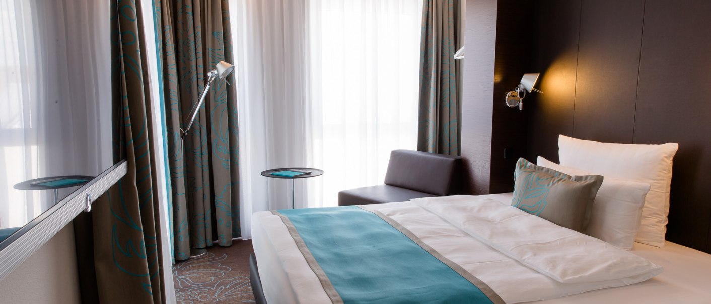 Modernes Hotelzimmer mit Doppelbett, blauen Akzenten, Sofa, Stehlampe und großen Fenstern mit Vorhängen., © Motel One Stuttgart-Bad Cannstatt