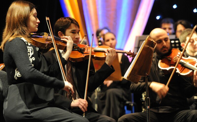 Ein Orchester spielt auf einer Bühne. Mehrere Musiker sind mit Geigen zu sehen, konzentriert auf ihre Instrumente. Hintergrund mit bunten Lichtern., © MCS Ein Orchester spielt auf einer Bühne. Mehrere Musiker sind mit Geigen zu sehen, konzentriert auf ihre Instrumente. Hintergrund mit bunten Lichtern., © MCS