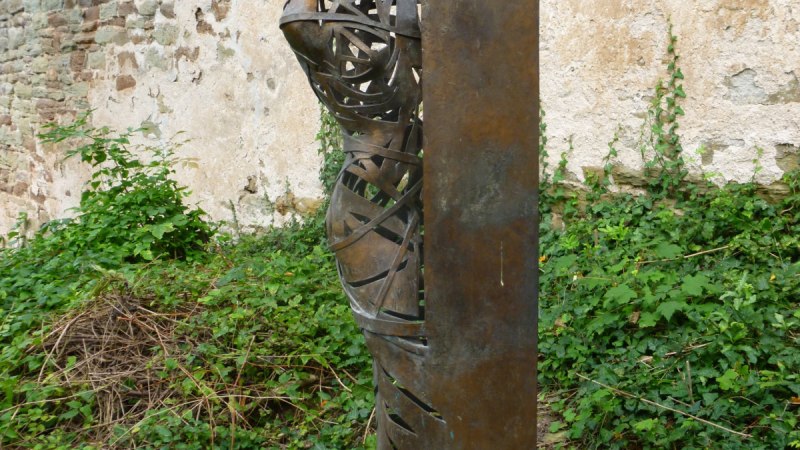 Metallskulptur eines Menschen, der hinter einer Wand hervorschaut, vor einer alten Steinmauer und umgeben von Pflanzen., © Natur.Nah. Schönbuch & Heckengäu Metallskulptur eines Menschen, der hinter einer Wand hervorschaut, vor einer alten Steinmauer und umgeben von Pflanzen., © Natur.Nah. Schönbuch & Heckengäu