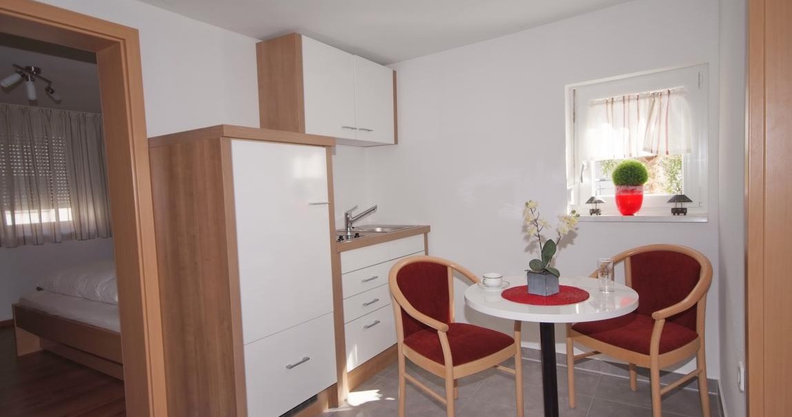 Kleine Küche mit weißem Schrank, Tisch und zwei roten Stühlen. Fenster mit Pflanze, daneben ein Schlafzimmer., © TOMAS