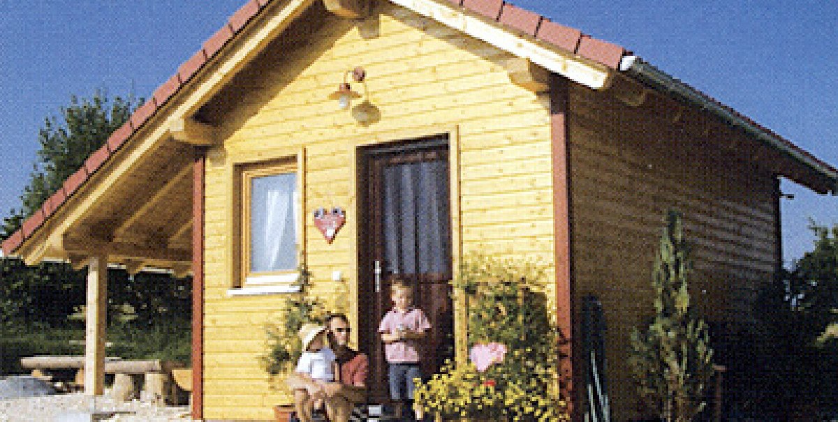 Kleines Holzhaus mit rotem Dach, davor sitzt eine Person mit Kind und Hund. Blumen und Pflanzen schmücken den Eingang. Der Himmel ist klar und blau.