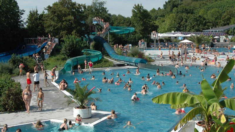 Ein belebtes Freibad mit zahlreichen Menschen im Wasser und auf den Rutschen. Umgeben von B&auml;umen und Pflanzen, herrscht eine sommerliche Atmosph&auml;re., &copy; Freibad Oberstenfeld