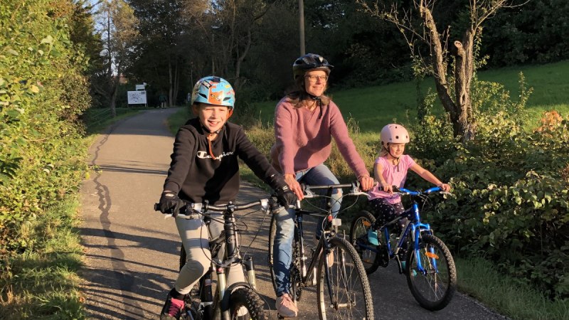 Drei Personen auf Fahrrädern fahren bei sonnigem Wetter auf einem Radweg durch eine grüne Landschaft., © T&M Drei Personen auf Fahrrädern fahren bei sonnigem Wetter auf einem Radweg durch eine grüne Landschaft., © T&M