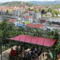 Panoramablick auf eine Stadt mit einem überdachten Sitzbereich im Vordergrund. Menschen sitzen an Tischen, im Hintergrund sind Gebäude und Hügel zu sehen., © Walter Laible