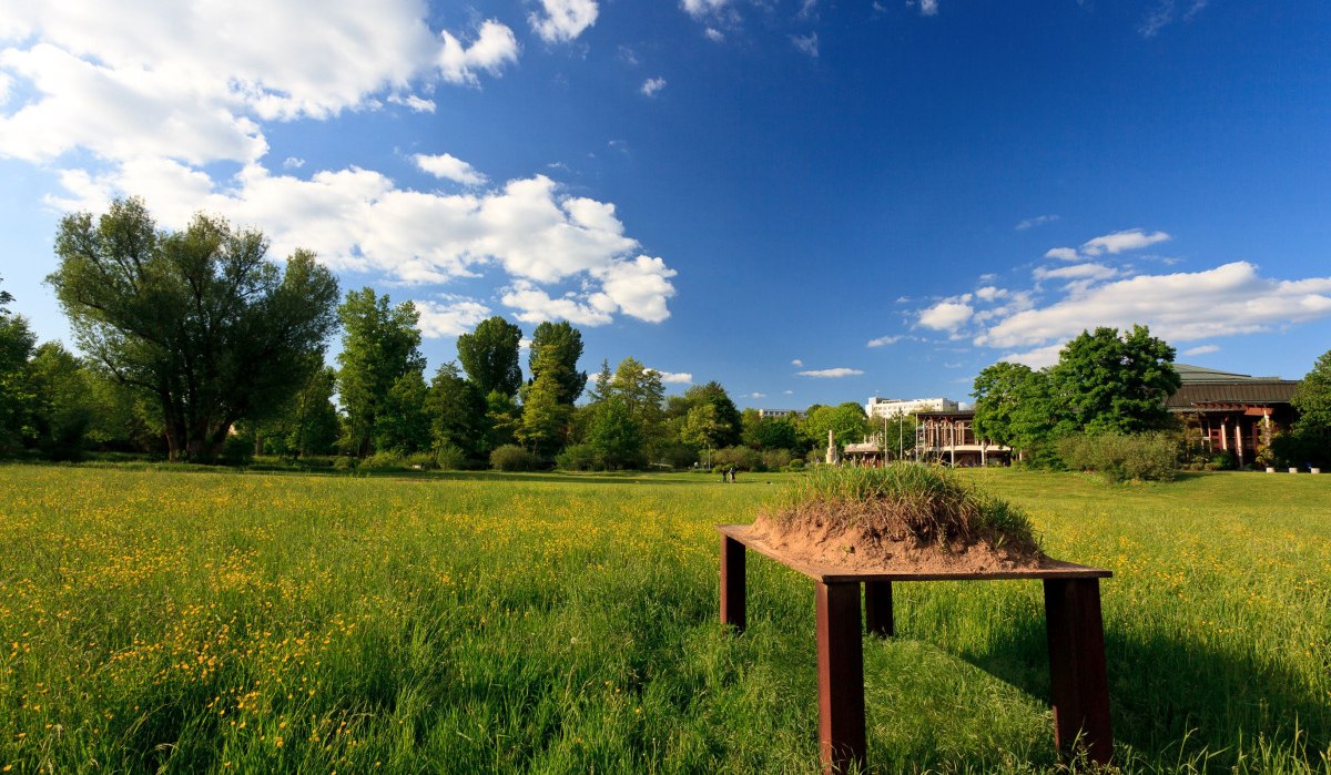 Ein Stahltisch mit Erdschollen steht auf einer grünen Wiese. Im Hintergrund sind Bäume und ein Gebäude zu sehen, der Himmel ist blau mit Wolken., © Remstal Tourismus e.V.
