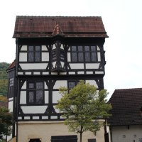 Ein Fachwerkhaus mit markantem Erker, daneben ein Kirchturm. Im Vordergrund ein Baum und eine Straßenlaterne.