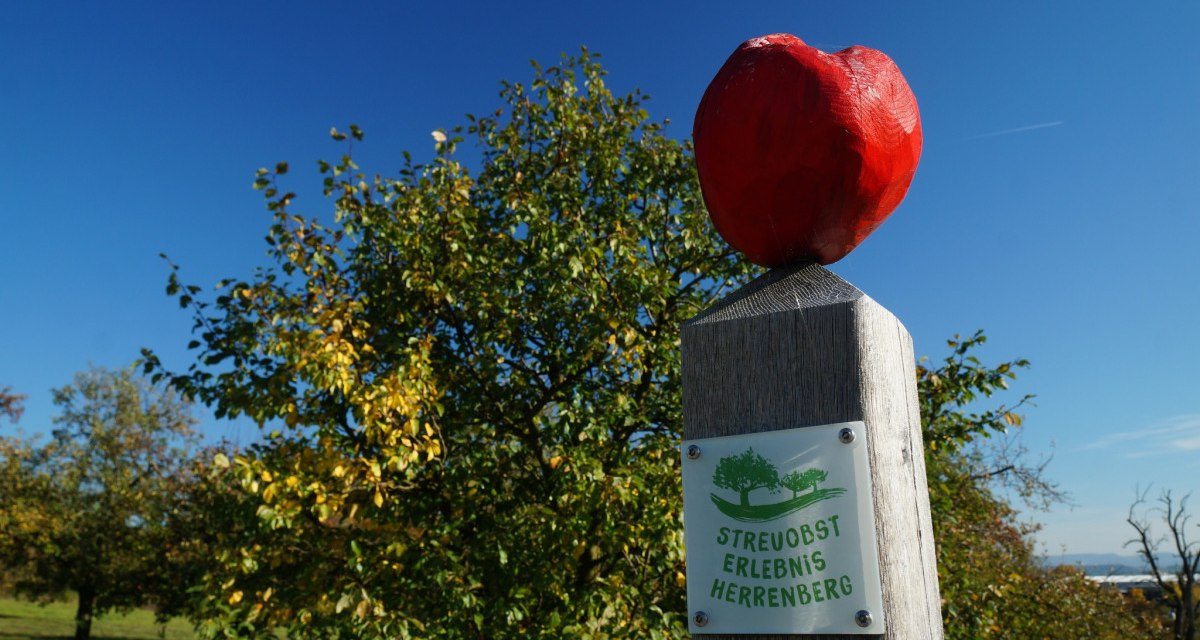 Ein Holzpfosten mit rotem Apfel und Schild 'Streuobst Erlebnis Herrenberg' vor blauem Himmel und B&auml;umen., &copy; Natur.Nah. Sch&ouml;nbuch & Heckeng&auml;u